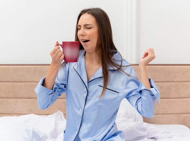 Young beautiful woman in blue pajamas sitting on bed with cup of coffee waking up yawning in bedroom interior on light background