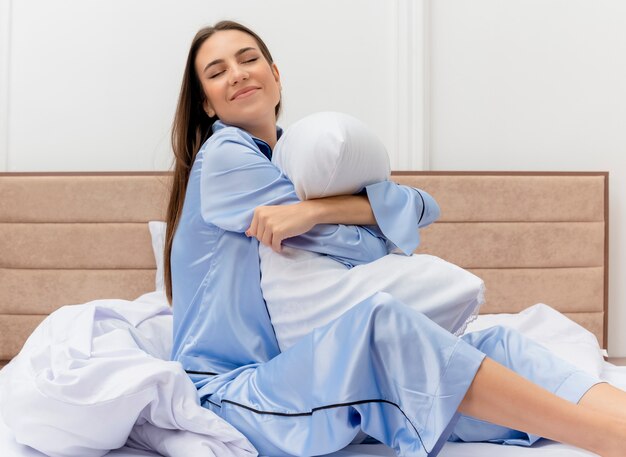 枕とベッドに座っている青いパジャマの若い美しい女性幸せで前向きに見える明るい背景の寝室のインテリアでうれしそうな笑顔
