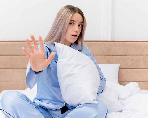 Young beautiful woman in blue pajamas sitting on bed with pillow lookign at camera making stop sing with hand in bedroom interior on light background