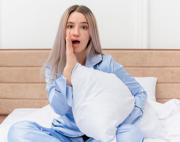 Young beautiful woman in blue pajamas sitting on bed with pillow looking at camera whispering a secret with hand near mouth in bedroom interior on light background