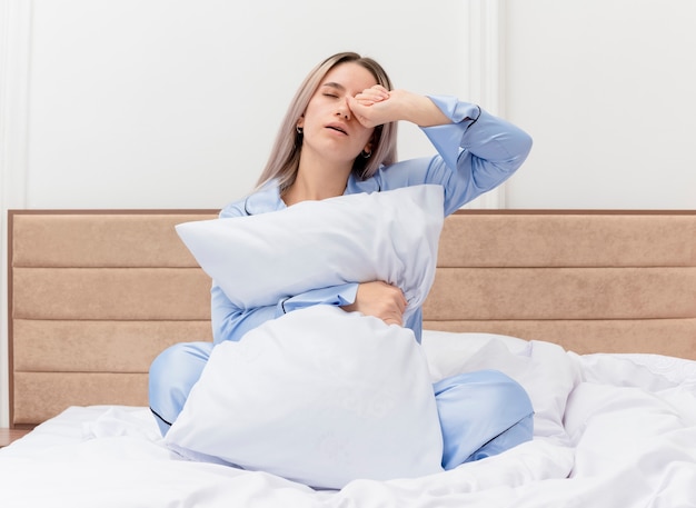 Молодая красивая женщина в синей пижаме сидит на кровати с подушкой, просыпаясь от ощущения утренней усталости в интерьере спальни