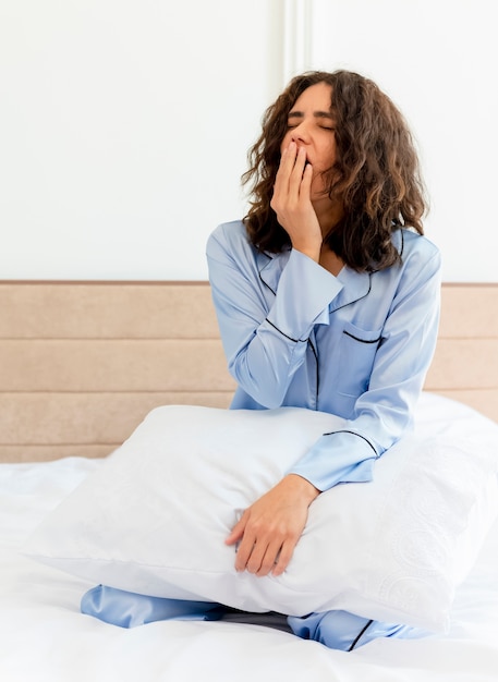 Молодая красивая женщина в синей пижаме, сидя на кровати с подушкой, просыпается, зевая в интерьере спальни на светлом фоне