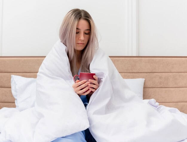 寝室のインテリアで気分が悪いコーヒーのカップを保持している毛布で包むベッドに座っている青いパジャマの若い美しい女性