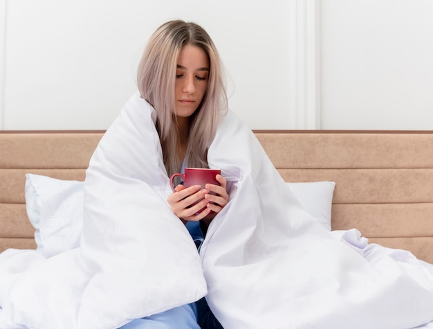 Young beautiful woman in blue pajamas sitting on bed wrapping in blanket holding cup of coffee feeling unwell in bedroom interior 