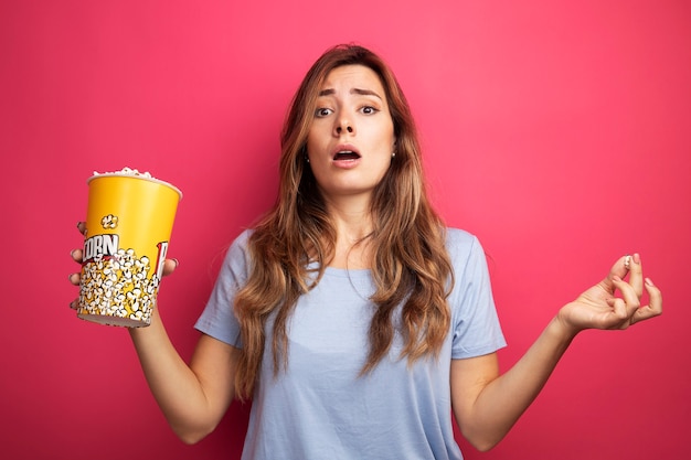 Young beautiful woman in blue t-shirt holding bucket with popcorn looking at camera surprised standing over pink background