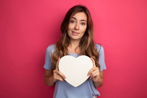 young beautiful woman in blue t-shirt holding cardboard heart looking at camera smiling cheerfully standing over pink background