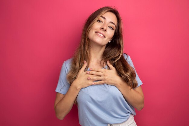 Young beautiful woman in blue t-shirt holding hands on her chest feeling thankful smiling friendly standing over pink background