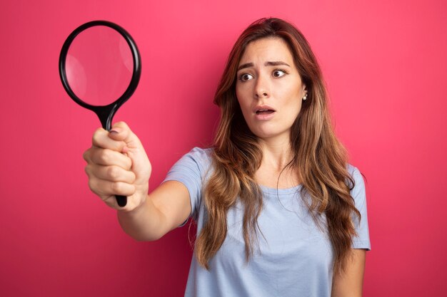 Young beautiful woman in blue t-shirt holding magnifying glass looking at it confused standing over pink background