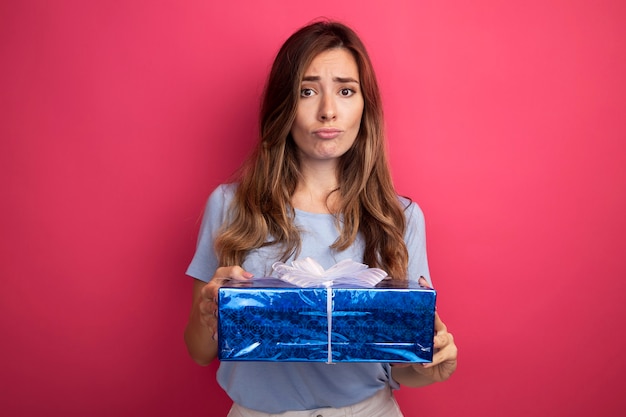 ピンクの背景の上に立って混乱しているカメラを見てプレゼントを保持している青いTシャツの若い美しい女性