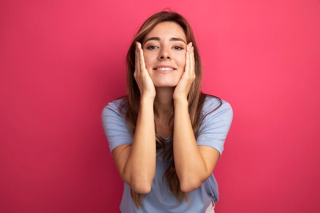 Giovane bella donna in maglietta blu che guarda la telecamera felice e positiva sorridente con le braccia sulle guance in piedi sul rosa