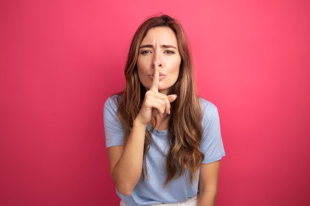 Young beautiful woman in blue t-shirt looking at camera making silence gesture with finger on lips standing over pink background