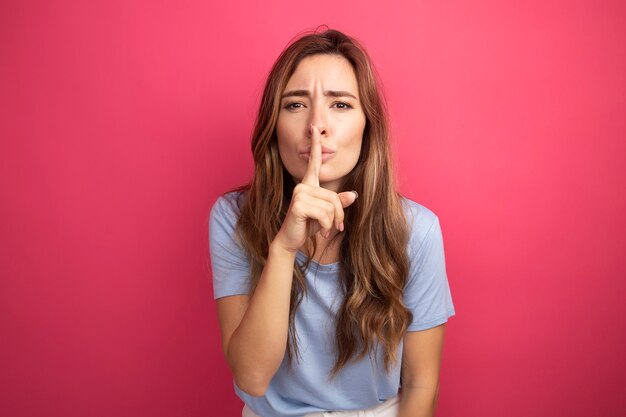 Молодая красивая женщина в синей футболке смотрит в камеру, делая жест молчания пальцем на губах, стоя на розовом фоне