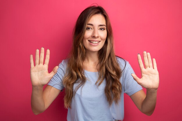 ピンクの上に立っている開いた手のひらを示す笑顔のカメラを見て青いTシャツの若い美しい女性