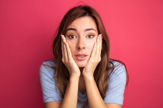 Young beautiful woman in blue t-shirt looking at camera surprised with hands on her cheeks standing over pink