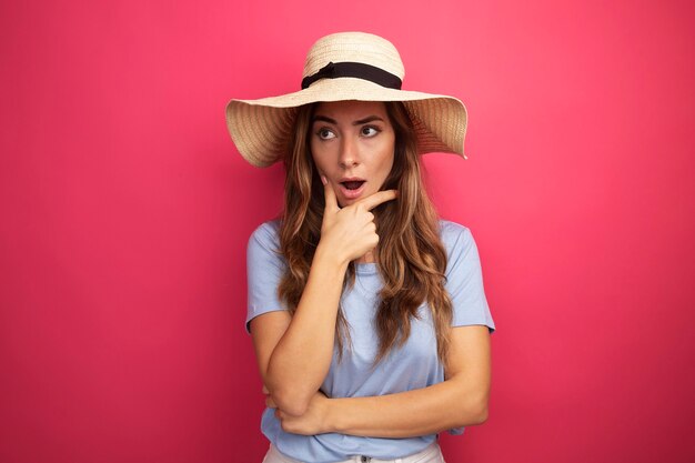 Молодая красивая женщина в синей футболке и летней шляпе смотрит в сторону удивленно, стоя на розовом фоне