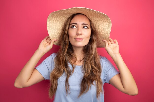 Young beautiful woman in blue t-shirt and summer hat looking aside with smile on face standing over pink background