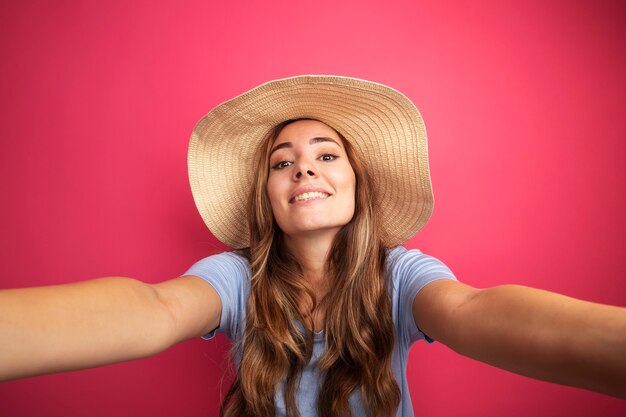 Young beautiful woman in blue t-shirt and summer hat looking at camera smiling cheerfully making welcoming gesture 