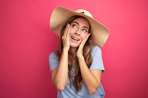Молодая красивая женщина в синей футболке и летней шляпе смотрит вверх счастливой и позитивной улыбкой