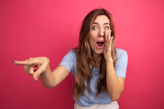 Young beautiful woman in blue t-shirt telling a secret with hand near mouth pointing with index finger at something standing over pink background