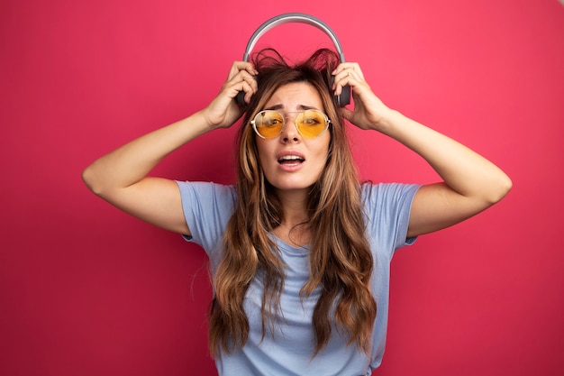 Foto gratuita giovane bella donna in maglietta blu che indossa occhiali gialli con le cuffie che guarda l'obbiettivo confuso e deluso