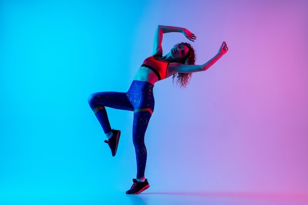 Young beautiful woman in bright sportwear isolated on gradient pink-blue background in neon light.