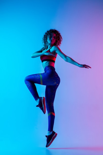 Young beautiful woman in bright sportwear isolated on gradient pink-blue background in neon light.