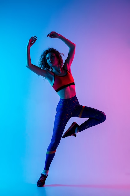 Young beautiful woman in bright sportwear isolated on gradient pink-blue background in neon light.