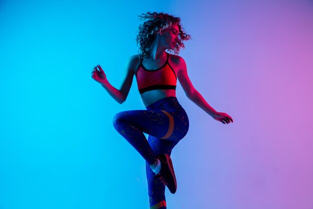 Young beautiful woman in bright sportwear isolated on gradient pink-blue background in neon light.