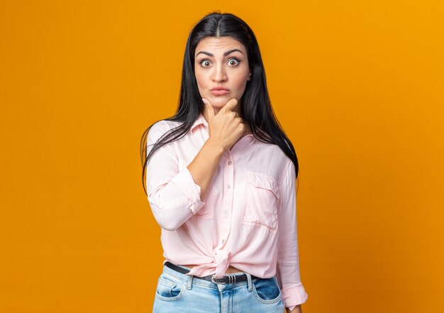 Young beautiful woman in casual clothes  confused and surprised standing over orange wall