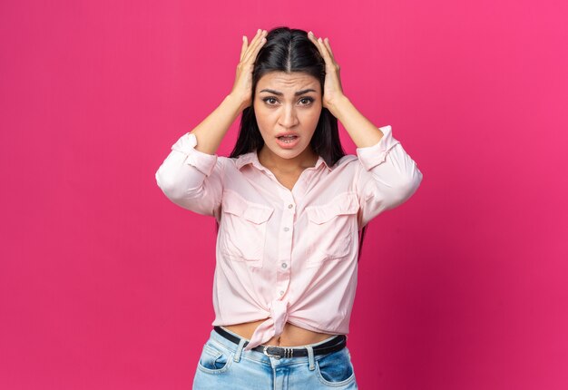 Young beautiful woman in casual clothes  confused and very anxious with hands on her head standing over pink wall