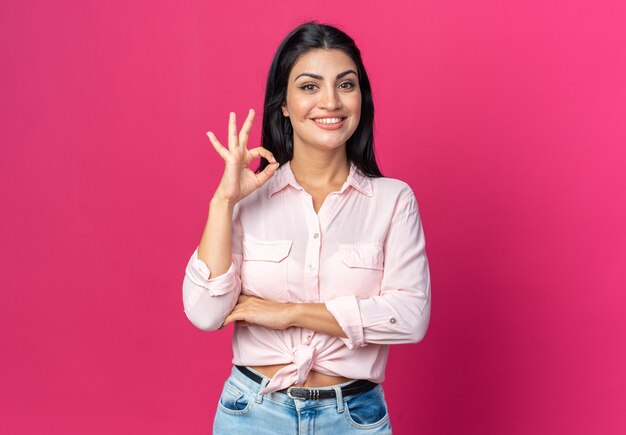 Young beautiful woman in casual clothes  happy and positive smiling cheerfully showing ok sign standing over pink wall