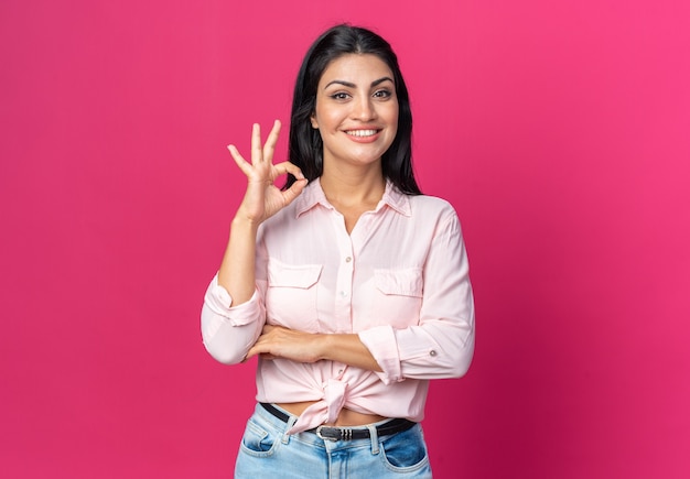 Young beautiful woman in casual clothes  happy and positive smiling cheerfully showing ok sign standing over pink wall