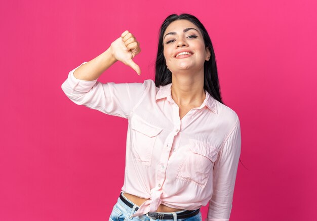 Young beautiful woman in casual clothes  happy and positive smiling cheerfully showing thumbs down standing over pink wall