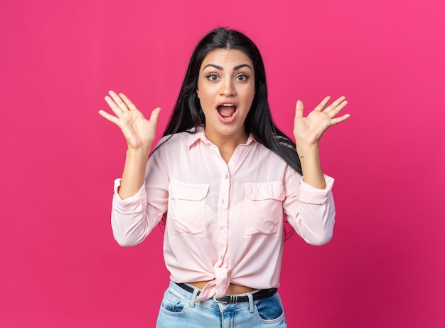 Young beautiful woman in casual clothes  happy and surprised with arms raised standing over pink wall