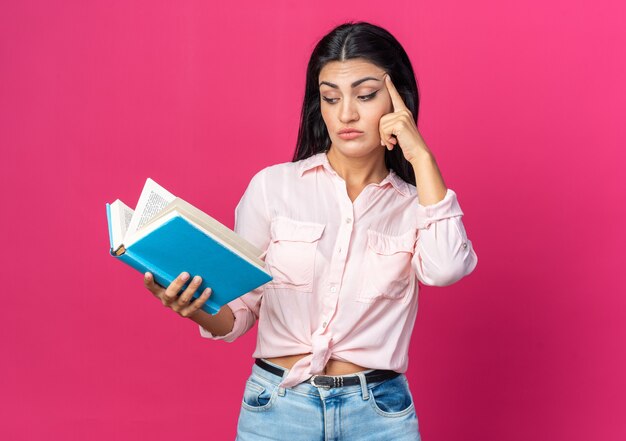 Young beautiful woman in casual clothes holding book looking at it puzzled with finger on temple standing on pink