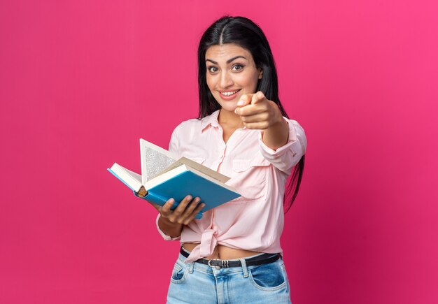 Молодая красивая женщина в повседневной одежде держит книгу, весело улыбаясь и позитивно указывая указательным пальцем на розовую стену