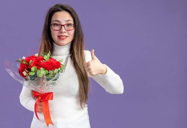 young beautiful woman in casual clothes holding bouquet of red roses looking at camera happy and positiveshowing thumbs up valentines day concept standing over purple wall