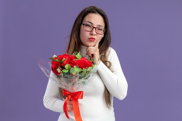 紫色の背景の上に立っている顔のバレンタインデーのコンセプトに物思いにふける表情でカメラを見て赤いバラの花束を保持しているカジュアルな服を着た若い美しい女性