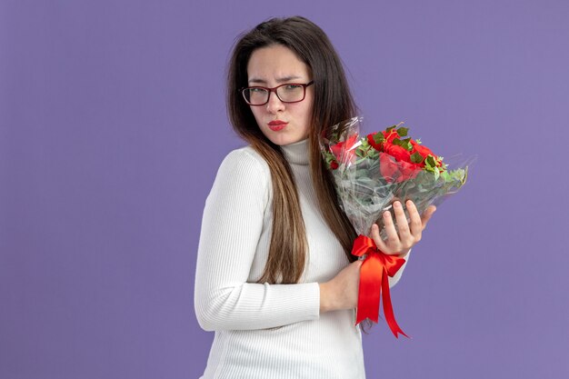 보라색 배경 위에 회의적인 식 발렌타인 데이 개념 서와 함께 카메라를보고 빨간 장미 꽃다발을 들고 캐주얼 옷에 젊은 아름 다운 여자