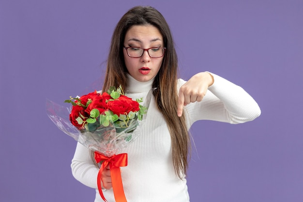 young beautiful woman in casual clothes holding bouquet of red roses looking down pointing with index finger down valentines day concept standing over purple background