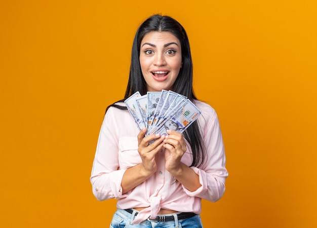 Young beautiful woman in casual clothes holding cash  happy and positive smiling cheerfully standing over orange wall