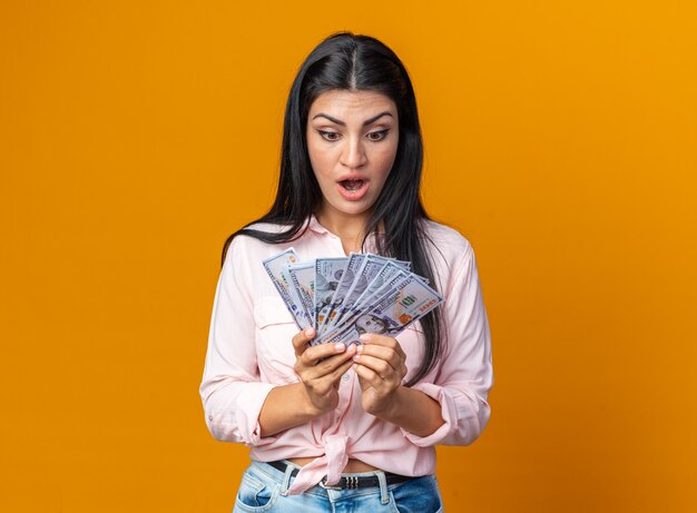 Young beautiful woman in casual clothes holding cash looking at money amazed and surprised