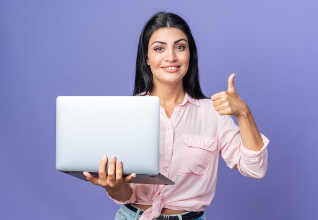 Молодая красивая женщина в повседневной одежде, держащая ноутбук, счастлива и позитивно смотрит, улыбается, уверенно показывает палец вверх