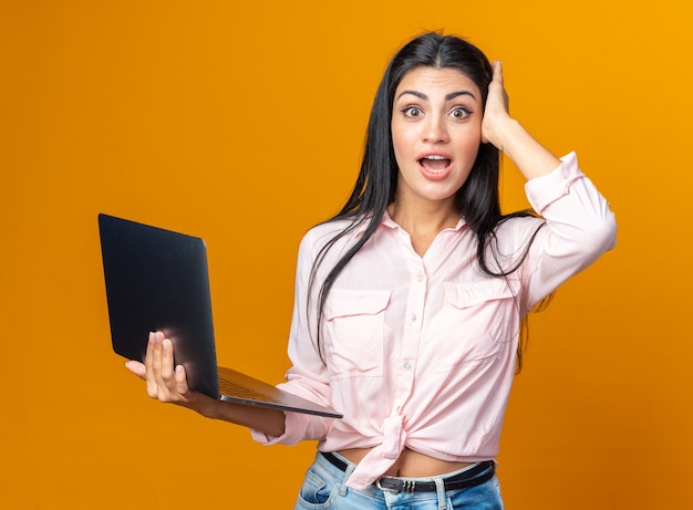 Young beautiful woman in casual clothes holding laptop looking at front happy and surprised standing over orange wall