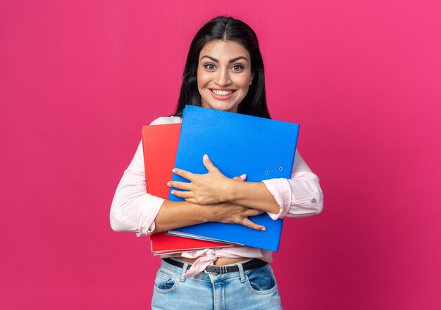 Young beautiful woman in casual clothes holding office folders looking at front happy and cheerful smiling broadly standing over pink wall