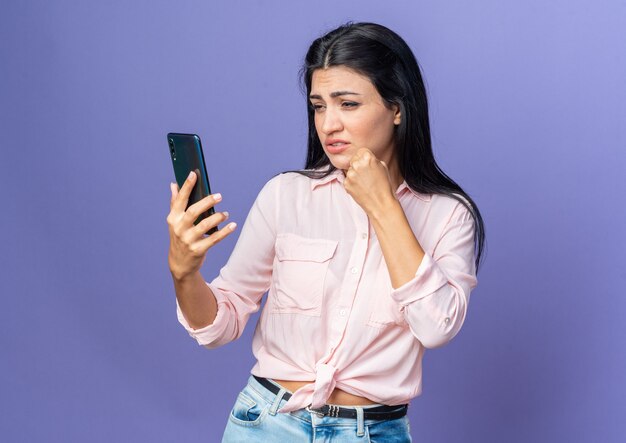 Young beautiful woman in casual clothes holding smartphone looking at screen confused and displeased