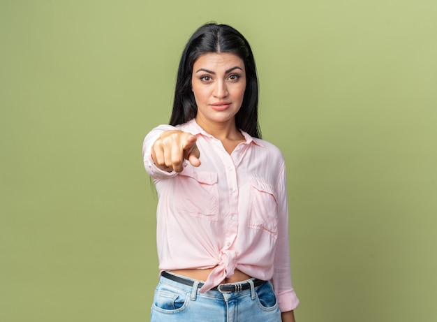 Young beautiful woman in casual clothes looking confident pointing with index finger  standing over green wall