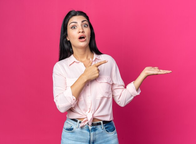 Young beautiful woman in casual clothes surprised pointing with index finger to the side presenting something with arm of her hand standing on pink