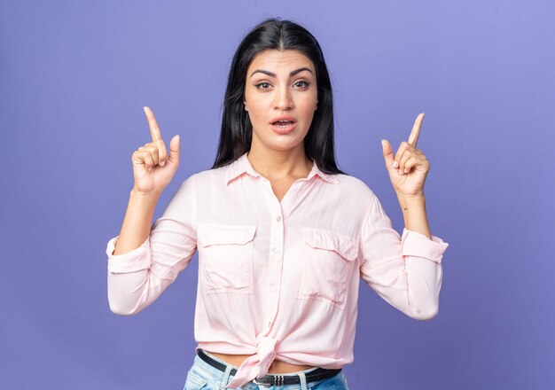 Young beautiful woman in casual clothes  surprised pointing with index fingers up standing over blue wall