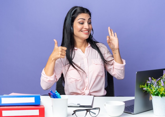 Giovane bella donna in abiti casual che indossa l'auricolare con microfono felice e allegro che mostra pollice in su facendo segno ok seduto al tavolo con il computer portatile sul muro blu che lavora in ufficio